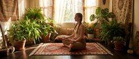 Woman in meditation pose sitting on yoga mat surrounded by potted plants in sunlit room with windows and curtains.