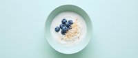 Overhead view of creamy yogurt bowl topped with fresh blueberries and granola on mint background, suitable for food blogs and wellness content.