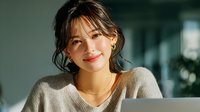 Young Asian businesswoman smiling while working on laptop in modern office environment. Professional female entrepreneur in casual sweater during productive workday.