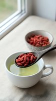 Dried goji berries on wooden spoon over green tea cup with bowl of superfood berries on windowsill for healthy nutrition and wellness lifestyle.