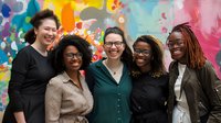 Five diverse professional women smiling together in modern office with colorful abstract art wall, representing teamwork and workplace collaboration.
