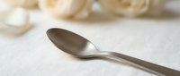 Stainless steel spoon on white linen fabric with blurred floral elements in background, suitable for dining, tableware, and culinary content.