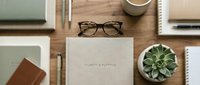 Organized workspace flatlay with eyeglasses, notebook, succulent plant, coffee cup, and stationery on wooden desk for business and productivity themes.