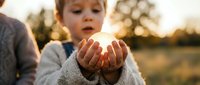 Young Caucasian child in cozy sweater cupping hands around glowing light at golden hour sunset, creating magical moment outdoors with warm bokeh background.