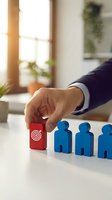 Businessman hand placing red target block next to blue wooden figures representing strategic planning and team leadership in modern office environment.
