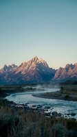 Majestic mountain peaks rise above a misty river valley at dawn, with rugged terrain and sparse vegetation in the foreground, ideal for travel and nature projects.