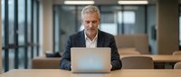 Middle-aged Caucasian male professional in business casual attire working on laptop at wooden desk in modern office environment with glass partitions and natural lighting.