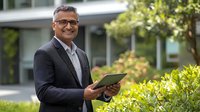 Middle-aged South Asian businessman in dark suit smiling while holding digital tablet outdoors against green garden background, representing success and leadership.