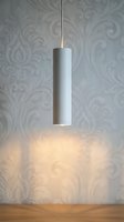 Minimalist white cylindrical pendant lamp hanging above wooden surface, casting warm glow on ornate damask wallpaper backdrop, contemporary interior lighting design for home decor and residential spaces.