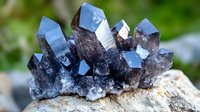 Smoky quartz crystal cluster formation with translucent blue tones on natural stone base against blurred green background, showing geometric crystalline structure.