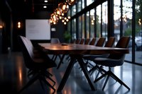 Modern conference room with wooden table and leather chairs illuminated by warm pendant lights, featuring floor-to-ceiling windows and moody atmosphere.
