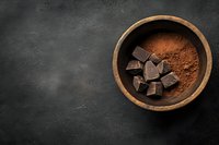 Dark chocolate pieces and cocoa powder in wooden bowl on black stone background, top view with copy space for dessert and confectionery designs.