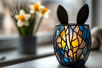 Decorative mosaic candle holder with bunny ears silhouette casting warm glow, next to spring daffodils on windowsill, creating cozy home atmosphere.