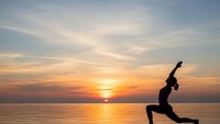 Woman practicing yoga warrior pose silhouette against golden sunset over calm ocean water creating peaceful meditation scene for wellness and mindfulness.