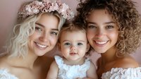 Diverse young women and baby girl wearing white lace and floral crown pose together with bright genuine smiles against neutral background, representing family joy and unity.