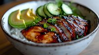 Grilled teriyaki chicken rice bowl with sliced avocado and fresh cucumber. Glazed meat topped with green onions served on steamed white rice in ceramic bowl.