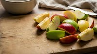 Fresh sliced apples in red, green and yellow varieties arranged on rustic wooden cutting board for healthy snacking and cooking preparation.