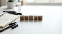 Wooden letter blocks spelling NOISE on white desk with notebook and pens in bright office workspace for communication and sound concept design.