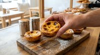 Hand holding freshly baked Portuguese egg tart on wooden board in bakery kitchen with golden custard filling and caramelized top ready to serve.