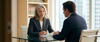 Professional blonde businesswoman in suit meeting with male colleague at modern office table discussing business strategy and corporate planning.