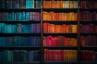 Colorful vintage library bookshelf with books arranged by color spectrum from blue to orange and red, creating a rainbow gradient effect in a dark room.