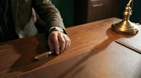 Mature male hand signing legal document at wooden desk with brass lamp and leather portfolio in office setting.