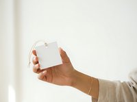 Unisex hand holding blank white square tag with string against neutral background for branding and labeling purposes.