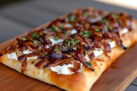 Homemade flatbread topped with caramelized onions, fresh herbs, and dollops of creamy goat cheese served on wooden cutting board, close up view of gourmet appetizer.
