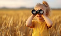 Curious blonde toddler girl in yellow sweater looking through binoculars in golden wheat field during sunset, exploring nature and adventure concept.