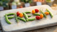 Fresh vegetables spelling the word FRESH on a rustic wooden cutting board, with green peas, cherry tomatoes, and rosemary sprigs.
