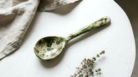 Handcrafted ceramic spoon with natural green glaze and organic texture on white marble surface with dried herbs for rustic kitchen and dining decor.