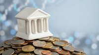 Miniature bank building model sitting on pile of coins with blurred financial chart background representing banking, savings, investment and economic growth concepts.