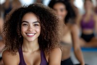 Young mixed-race woman with curly hair smiling during fitness class workout session in gym with diverse group of people exercising in background.