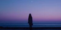 Silhouette of woman standing on beach at twilight, gazing at purple and blue horizon with crescent moon in peaceful solitude.