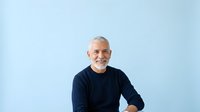 Senior man with white hair and beard, Caucasian ethnicity, wearing dark navy sweater, smiling at camera against light blue background. Suitable for healthcare, wellness, lifestyle and corporate projects.