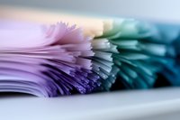 Macro shot of colorful paper sheets stacked in gradient from pink to turquoise, creating abstract wave pattern with soft focus and artistic blur effect.