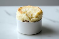 Homemade shepherd's pie in white ramekin with golden brown mashed potato topping, traditional comfort food against light background, close up view.