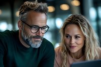 Mature Caucasian couple looking at digital tablet together, man with gray beard wearing glasses and green sweater, woman with blonde hair smiling warmly.