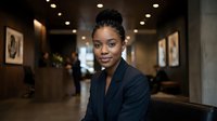 Professional African American businesswoman in dark suit smiling confidently in modern corporate office lobby with warm lighting and contemporary decor.