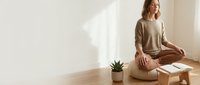 Young woman practicing mindfulness meditation sitting cross-legged on wooden floor in bright minimalist room with natural sunlight and small plant.