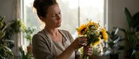 Middle-aged Caucasian woman arranging fresh sunflower bouquet in bright home interior with natural light and houseplants creating cozy domestic atmosphere.