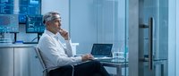 Middle-aged Caucasian male professional in business casual attire working on laptop in modern glass office, analyzing data on screen with thoughtful expression.