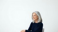 Mature woman with long gray hair and fair complexion wearing dark navy sweater, seated against white background for corporate portraits and professional headshots.