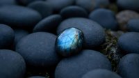 Blue labradorite gemstone with iridescent flash surrounded by smooth dark river rocks creating mystical mineral composition for jewelry design.