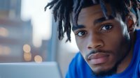 Young African American man with dreadlocks in blue hoodie looking thoughtfully at camera with urban city lights bokeh background at dusk.