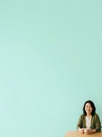 Smiling Asian woman in olive cardigan sitting at wooden table against mint green background, holding coffee cup, professional portrait for business and lifestyle use.
