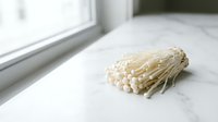 Enoki mushrooms with thin delicate stems and small caps arranged on white marble surface near window, suitable for food photography and culinary content.