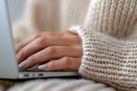 Female hands typing on laptop keyboard wearing cozy knit sweater. Soft focus close up of manicured fingers working from home in warm atmosphere.