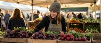 Young blonde woman in knit hat and apron selecting fresh beets at outdoor farmers market with wooden crates full of colorful root vegetables and shoppers.