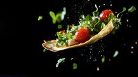 Levitating taco shell with fresh tomatoes, green herbs and spices captured in dynamic motion against dark background with water droplets and flying ingredients.